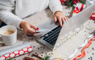Woman buying presents online with silver credit card and laptop computer, drinks coffee among gift boxes.
