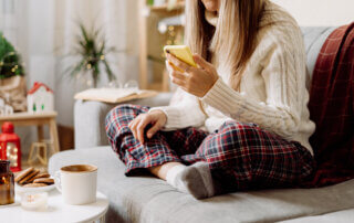 Cozy woman in knitted winter warm socks, sweater and checkered plaid with phone, drinking hot cocoa or coffee in mug, during resting on couch at home.