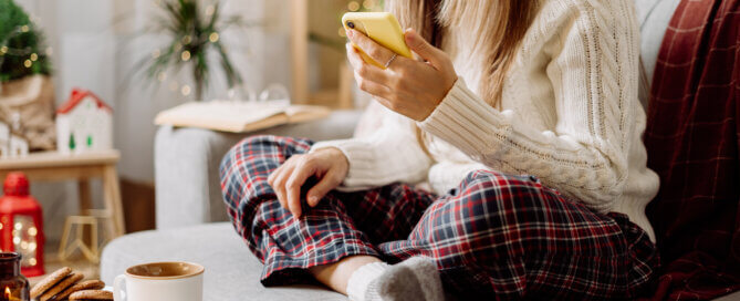 Cozy woman in knitted winter warm socks, sweater and checkered plaid with phone, drinking hot cocoa or coffee in mug, during resting on couch at home.